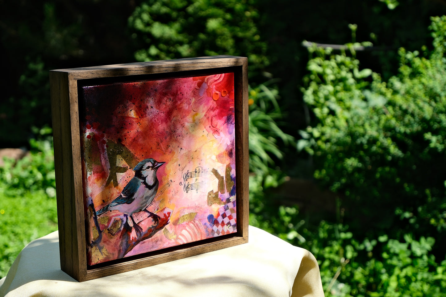 Framed artwork with a colorful abstract design and a bird illustration, placed outdoors on a green background.