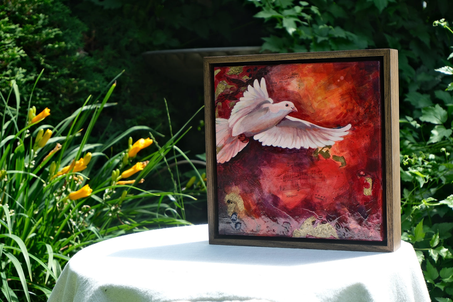Framed artwork of a dove on a table outdoors with greenery and flowers in the background