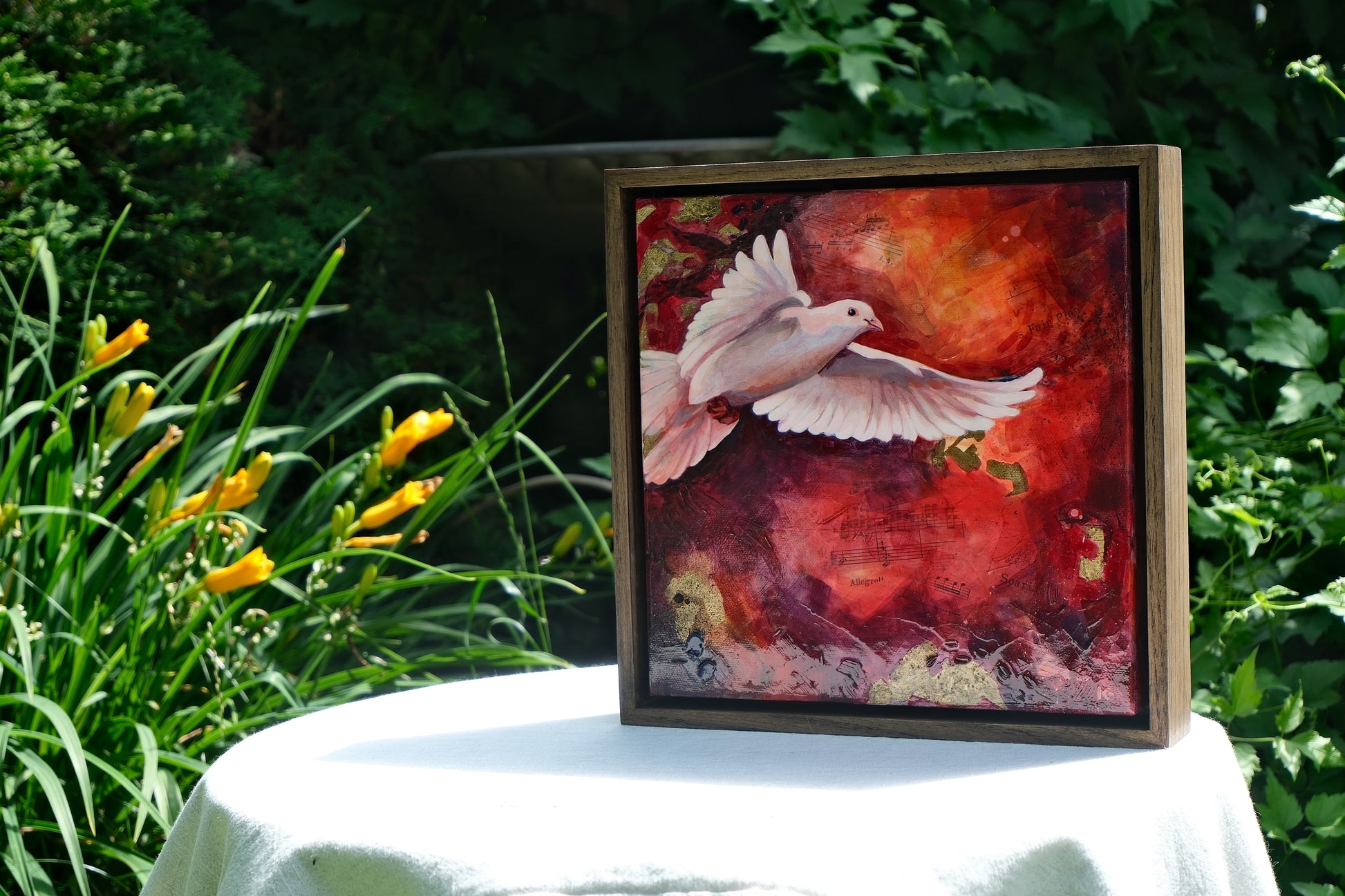 Framed artwork of a dove on a table outdoors with greenery and flowers in the background